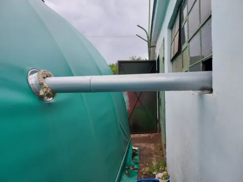 Rura doprowadza ciepłe powietrze do zielonej hali namiotowej. Poszycie jest zaoblone co wskazuje na łukową konstrukcję hali.