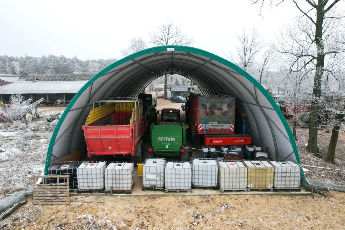 Łukowa wiata na gospodarstwie w zimowej scenerii; wewnątrz stoją ciągniki i maszyny rolnicze, z przodu ustawione paletopojemniki IBC. Zadaszenie chroni sprzęt przed śniegiem i deszczem, ułatwiając organizację placu.