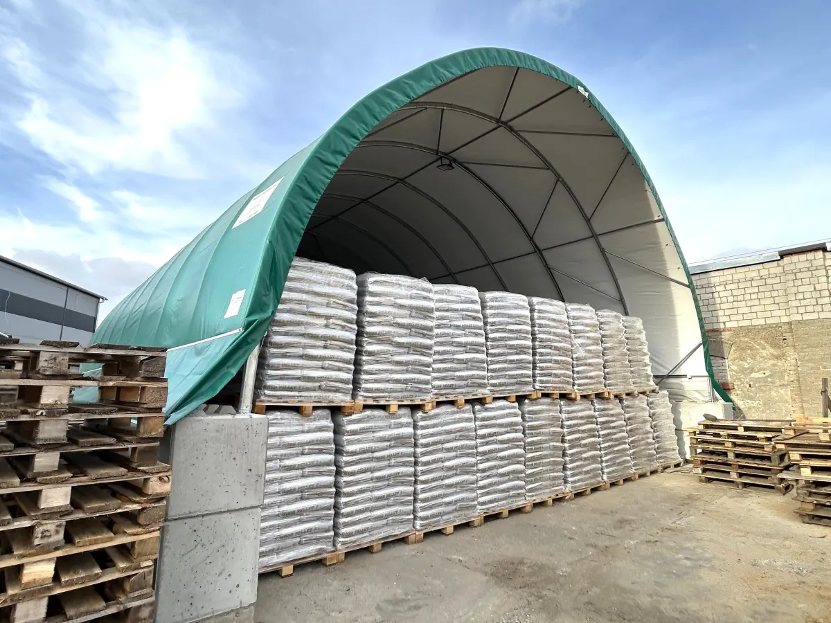 Pod wiatą łukową składowane są szczelnie ułożone palety z towarem zapakowanym w worki. Zadaszenie chroni ładunek, a front pozostaje otwarty dla wózków.