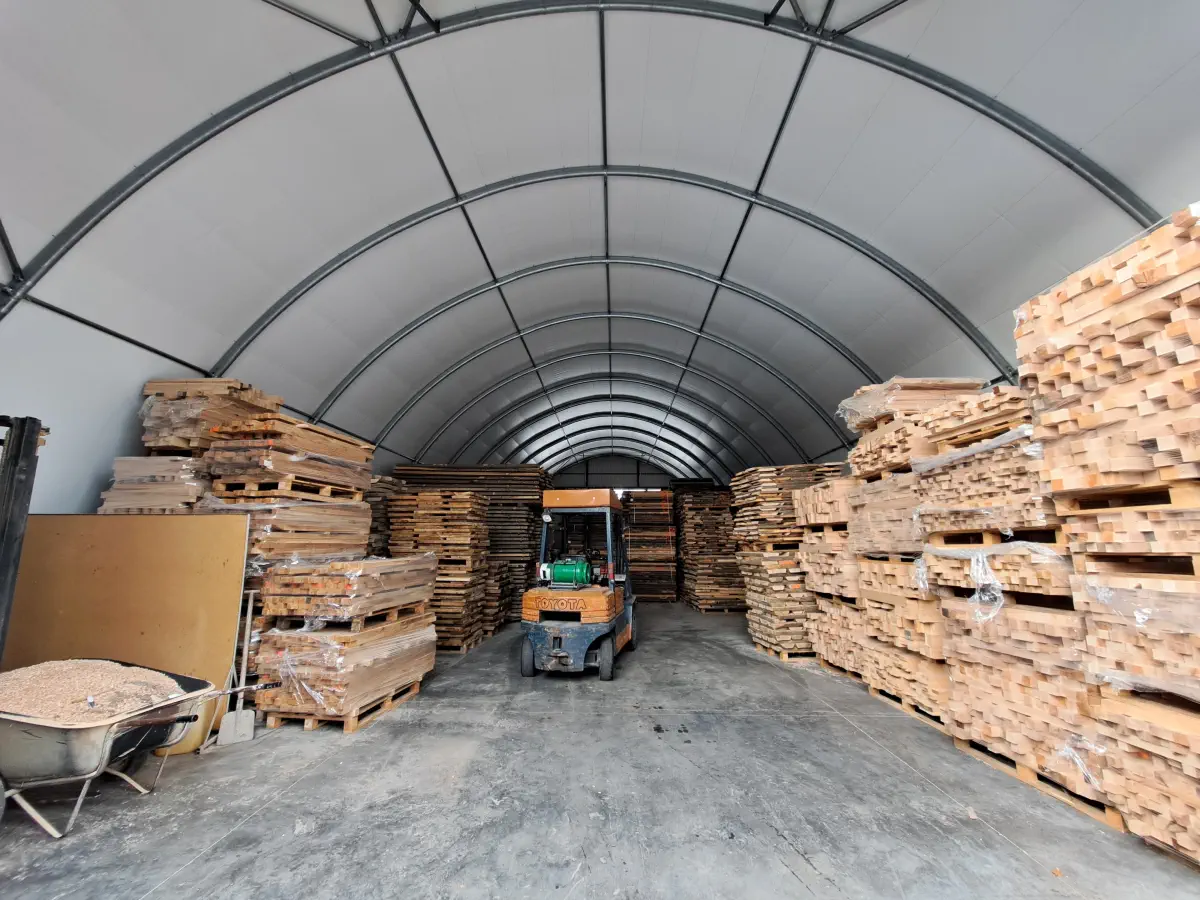 W środku łukowej hali widać uporządkowane stosy desek na paletach oraz wózek widłowy. Gładka posadzka i wysoki prześwit ułatwiają składowanie i logistykę.