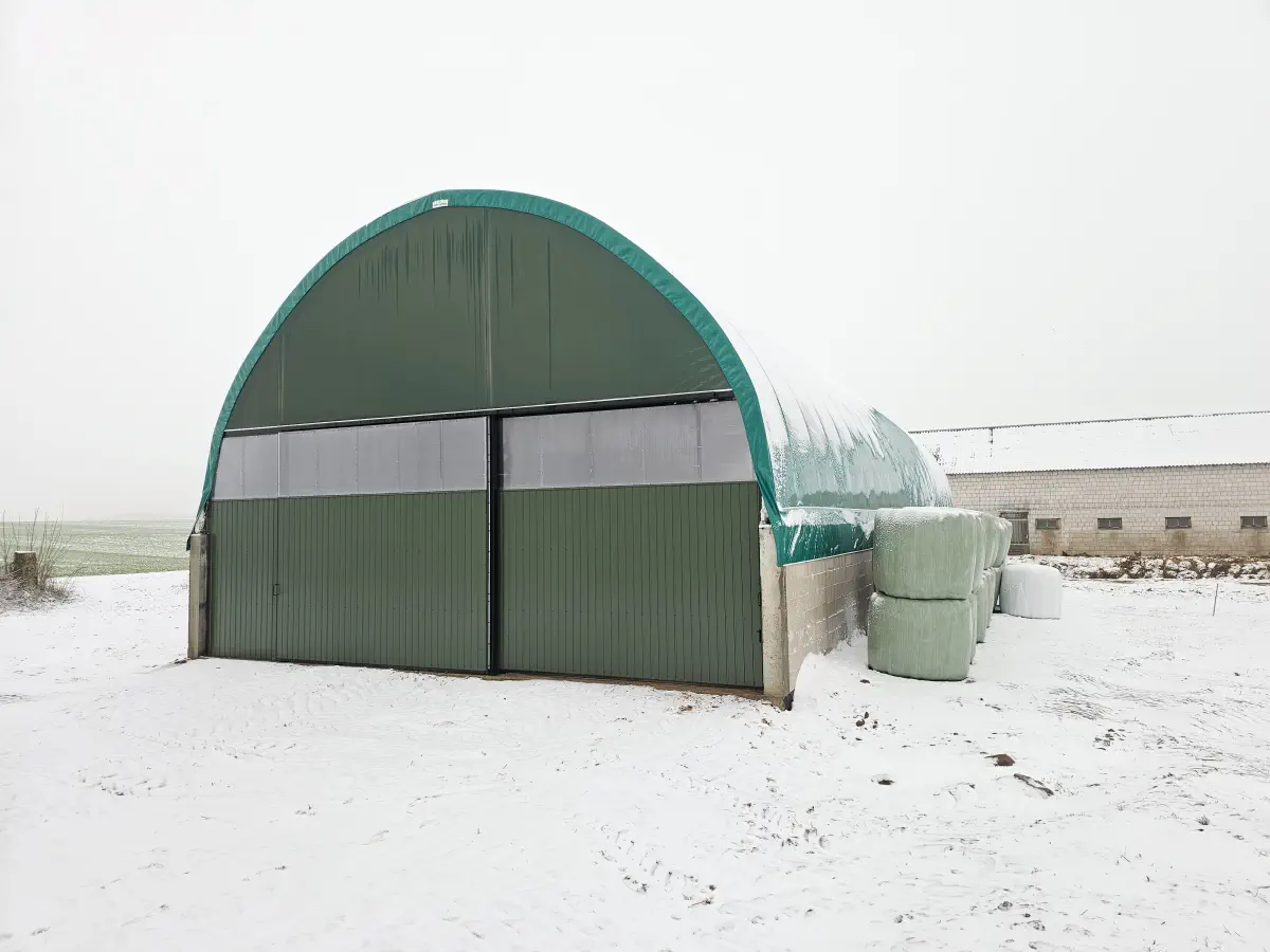 Zamknięta hala z przesuwnymi wrotami i bocznymi murkami z bloczków, pokryta warstwą śniegu. Obok ułożone bele sianokiszonki; obiekt służy do całorocznego magazynowania w gospodarstwie.