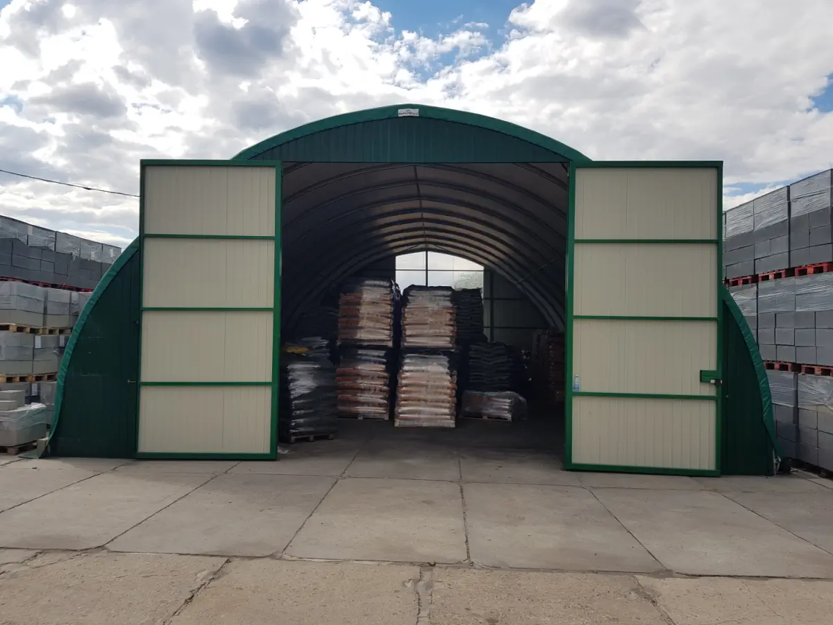 Hala łukowa z otwartymi, przesuwnymi drzwiami; w środku widać palety z towarem. Obiekt stoi na utwardzonym placu pomiędzy składowanymi materiałami.