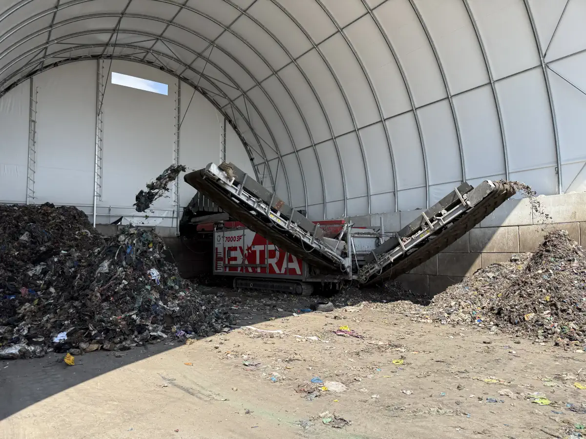 Wnetrze hali namiotowej do sortowania odpadów. Wewnątrz widać składowisko odpadów oraz maszynę, która rozdziela odpady na dwa stosy.