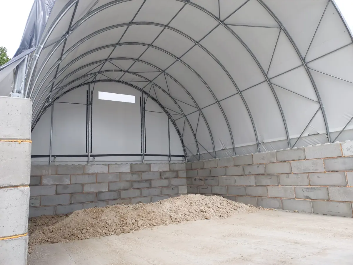 Zdjęcie od wewnątrz pokazujące stalową konstrukcję zadaszenia osadów pokrytą jasną plandeką. Na betonowej posadzce, pomiędzy wysokimi murami z bloków, znajduje się niewielka pryzma suchego osadu.