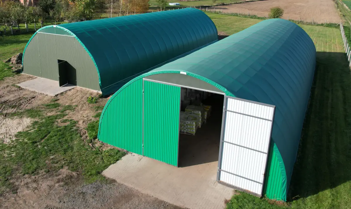 Dwie hale łukowe pokryte zieloną membraną PVC z okładziną z blachy trapezowej na ścianach szczytowych, wyposażone w drzwi wejściowe oraz otwartą bramę z widocznym paletyzowanym towarem wewnątrz. Konstrukcje posadowione na betonowych płytach fundamentowych tworzą funkcjonalny kompleks magazynowy dla gospodarstwa rolniczego.