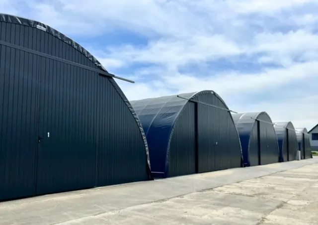 Kompleks hal łukowych ustawionych w rzędzie, z ścianami szczytowymi wykonanymi z grafitowych paneli stalowych trapezowych oraz szarą membraną PVC pokrywającą konstrukcję łukową. Obiekty magazynowe z drzwiami wejściowymi po lewej stronie każdej hali, połączone wspólną betonową drogą dojazdową umożliwiającą sprawny dostęp do poszczególnych modułów. Rozwiązanie modułowe pozwalające na elastyczne rozbudowanie przestrzeni magazynowej poprzez dodawanie kolejnych segmentów.