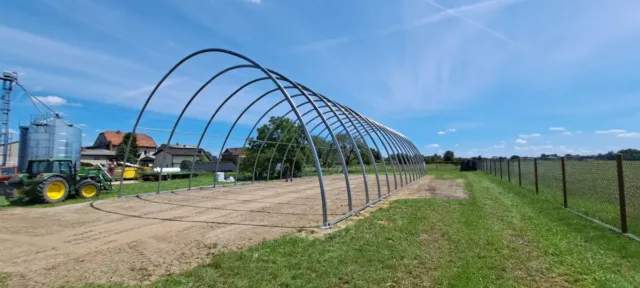 Stalowy szkielet hali namiotowej w trakcie montażu, składający się z szeregu łukowych ram metalowych zakotwiczonych w betonowym podłożu. Konstrukcja nośna przygotowana do okrycia membraną PVC, ustawiona na terenie gospodarstwa rolniczego obok budynków mieszkalnych i silosów zbożowych. Widoczny żółty traktor w tle oraz ogrodzenie sąsiedniej posesji, ilustrujące typowe zastosowanie hal namiotowych w rolnictwie.